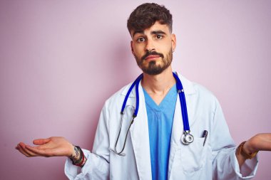Young doctor man with tattoo wearing stethocope standing over isolated pink background clueless and confused expression with arms and hands raised. Doubt concept. clipart