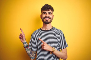 Çizgili t-shirt giyen dövme giyen genç adam izole sarı arka plan üzerinde gülümseyerek ayakta ve yan iki el ve parmakları ile işaret kamera bakıyor.