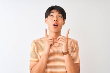 Izole beyaz arka plan üzerinde duran rahat çizgili t-shirt giyen genç Çinli adam şaşırttı ve yukarı bakarak ve parmakları ile işaret ve kolları kaldırdı şaşırttı.