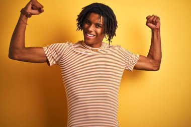 Çizgili t-shirt giyen dreadlocks ile Afro adam kolkasları gururla gülümseyen gösteren izole sarı arka plan üzerinde duruyor. Fitness konsepti.
