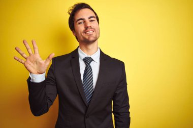 Takım elbise giyen ve kravat giyen genç yakışıklı işadamı izole sarı arka plan üzerinde duran gösteren ve parmakları ile işaret ederken emin ve mutlu gülümseyerek beş numara.