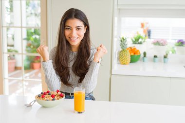 Sabahları sağlıklı kahvaltı yapan genç bir kadın şaşkınlık ve şaşkınlık içinde kollarını kaldırarak ve gözlerini açarak başarıyı kutluyor. Kazanan konsept.