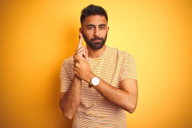 İzole sarı arka plan üzerinde duran t-shirt giyen genç hintli adam el hareketi ile sembolik silah holding, çekim silah öldürme oynarken, öfkeli yüz