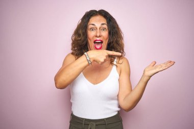 Middle age senior woman standing over pink isolated background amazed and smiling to the camera while presenting with hand and pointing with finger.