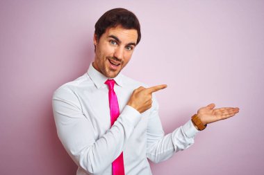 Genç yakışıklı işadamı gömlek ve kravat izole pembe arka plan üzerinde duran giyen hayran ve el ile sunarken kameraya gülümseyerek ve parmak ile işaret.