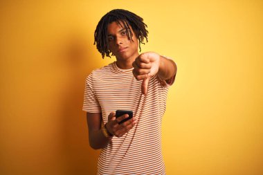 Kızgın yüz ile izole sarı arka plan üzerinde akıllı telefon kullanarak dreadlocks ile Dreadlocks ile Afro Amerikan adam, aşağı başparmak ile sevmediğim gösteren negatif işareti, ret kavramı