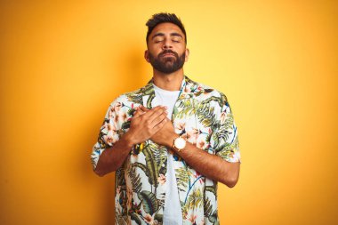 Tatilde genç hintli adam izole sarı arka plan üzerinde kapalı gözleri ve yüzünde minnettar jest ile göğüs üzerinde gülümseyerek yaz çiçek gömlek giyiyor. Sağlık kavramı.