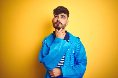 Yağmurluk giyen genç yakışıklı adam izole sarı arka plan üzerinde duran çene üzerinde soru, dalgın ifade düşünme. Düşünceli bir yüzle gülümsüyor. Şüphe kavramı.