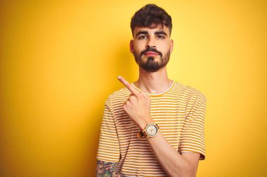 Çizgili t-shirt giyen dövme giyen genç adam izole sarı arka plan üzerinde duran yan reklam gösteren el parmağı ile işaret, ciddi ve sakin yüz