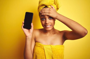 Duştan sonra havlu giyen afro kadın izole sarı arka plan üzerinde smatrphone gösteren baş el ile vurguladı, utanç ve sürpriz yüz ile şok, kızgın ve sinirli. Korku ve hata için üzgün.