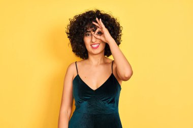 İzole sarı arka plan üzerinde zarif elbise giyen kıvırcık saçlı genç arap kadın el gülümseyerek ok jest yapıyor, mutlu yüz ile parmaklardan bakan göz.
