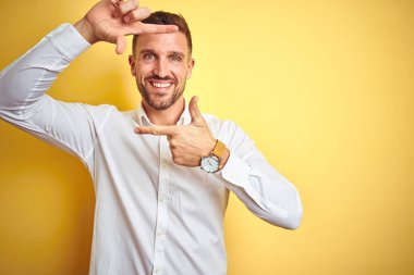 Sarı izole edilmiş arka planda zarif beyaz gömlek giyen yakışıklı bir adam gülümsüyor ve mutlu yüzlü elleri ve parmakları olan bir çerçeve yapıyor. Yaratıcılık ve fotoğrafçılık kavramı.