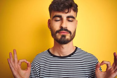 Çizgili t-shirt giyen dövme giyen genç adam izole sarı arka plan üzerinde duran rahatlayın ve gözleri parmakları ile meditasyon jest yapıyor kapalı gülümseyerek. Yoga konsepti.
