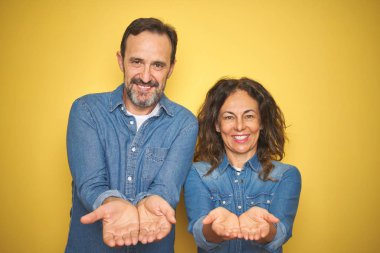 Güzel orta yaş çift birlikte izole sarı arka plan üzerinde duran birlikte alma veya jest veren avuç içi ile gülümseyerek. Tutma ve koruma