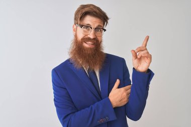 Genç kızıl İrlandalı işadamı yüzünde büyük bir gülümseme ile izole beyaz arka plan üzerinde takım elbise ve gözlük giyen, kameraya bakarak yan el ve parmak ile işaret.
