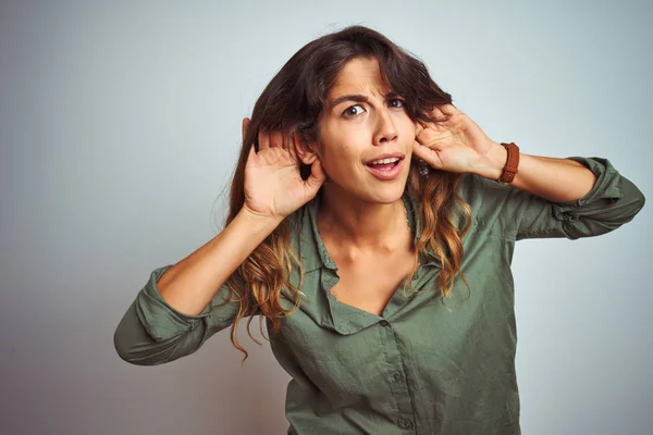 若い美しい女性の緑のシャツ灰色の隔離された背景の上に立って耳のジェスチャーで両方の手を聞くしようとすると ゴシップを好奇心 聴覚障害 聴覚障害 — ストック写真