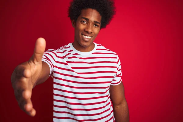 孤立した赤い背景の上にストライプのTシャツを着たアフロヘアの若いアフリカ系アメリカ人男性は 挨拶と歓迎として握手を提供する友好的な笑顔 ビジネスの成功 — ストック写真