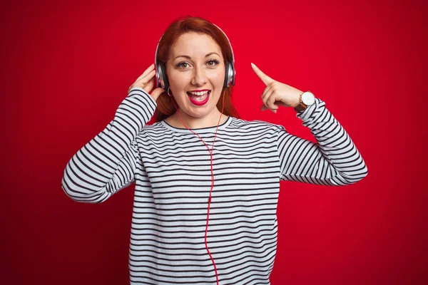 年轻的红头发女人在红色孤立的背景下通过耳机听音乐 她非常高兴地用手和手指指指点点 — 图库照片