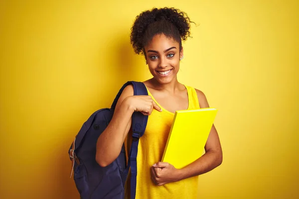 American student woman wearing backpack holding notebook over isolated yellow background with surprise face pointing finger to himself