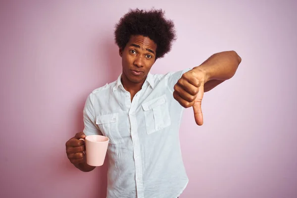 Young african american man drinking a cup of coffee standing over isolated pink background with angry face, negative sign showing dislike with thumbs down, rejection concept