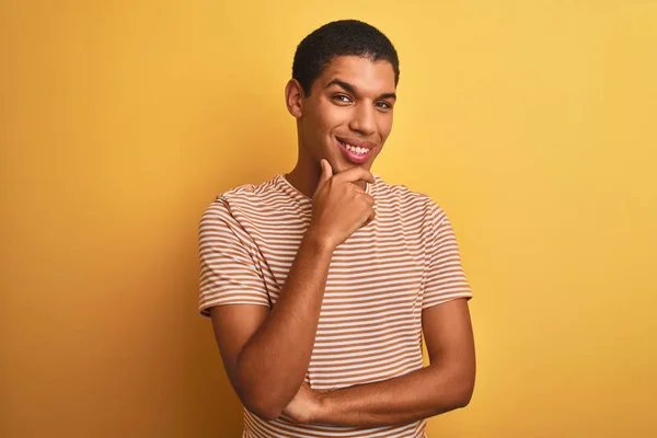 Young handsome arab man wearing striped t-shirt standing over isolated yellow background looking confident at the camera smiling with crossed arms and hand raised on chin. Thinking positive.