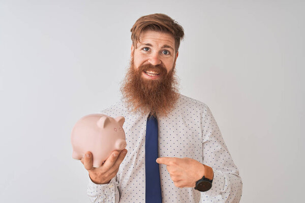 Young redhead irish businessman holding piggy bank over isolated white background with surprise face pointing finger to himself