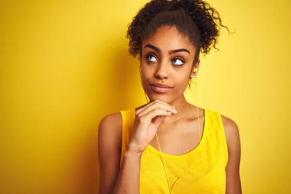 African american woman listening to music using earphones over isolated yellow background with hand on chin thinking about question, pensive expression. Smiling with thoughtful face. Doubt concept.