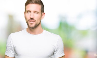 Casual beyaz t-shirt seksi bir ifadeyle, neşeli ve mutlu yüzünü kameraya bakarak göz kırpıyor giyen yakışıklı adam.