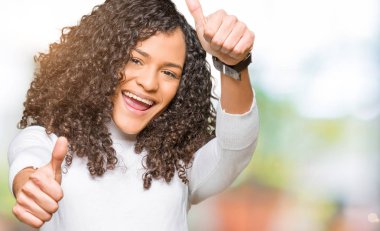 Kıvırcık saçlı Genç güzel kadın el ile olumlu jest yapıyor onaylamak balıkçı yaka kazak giyen, thumbs up gülümseyen ve başarı için mutlu. Kamera bakarak, kazanan jest.