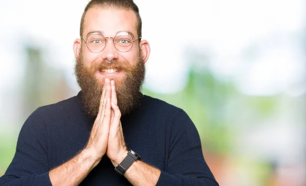 Young blond man wearing glasses and turtleneck sweater praying with hands together asking for forgiveness smiling confident.