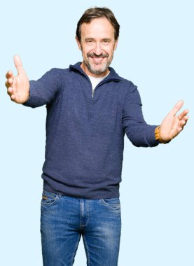 Orta yaş yakışıklı adam sarılmak için açık kollar ile gülümseyen kamera bakarak bir kazak giyiyor. Mutluluğu kucaklayan neşeli ifade.