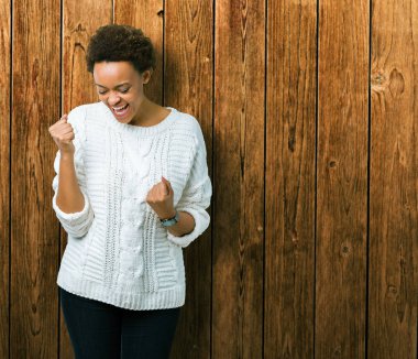 Üzerinde kazak giyen güzel genç Afro-Amerikan kadın gülümseyerek ve başarı için çığlık arka plan çok mutlu ve heyecanlı kaldırdı, silah ile kazanan jest yapıyor izole. Kutlama kavramı.