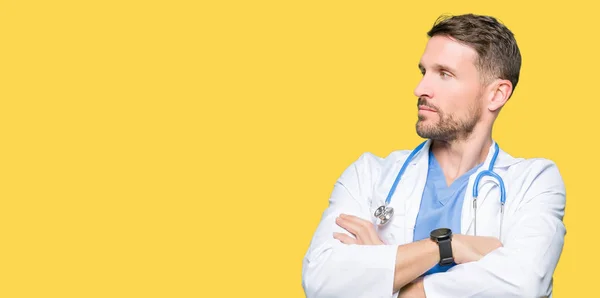 英俊的医生人穿着医疗制服在孤立的背景微笑着看着身边与双臂交叉的信心和自信 — 图库照片