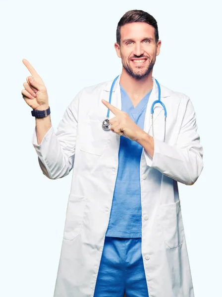英俊的医生男子穿着医疗制服在孤立的背景微笑着 看着相机指向镜头 用两只手和手指指着一边 — 图库照片
