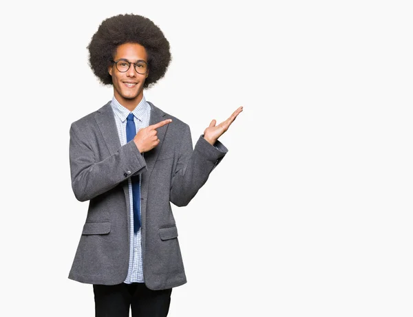 Young african american business man with afro hair wearing glasses amazed and smiling to the camera while presenting with hand and pointing with finger.