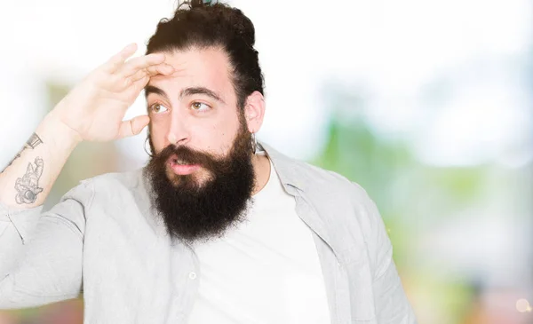 Young man with long hair, beard and earrings very happy and smiling looking far away with hand over head. Searching concept.