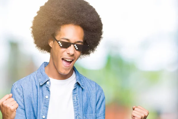 Young african american man with afro hair wearing thug life glasses very happy and excited doing winner gesture with arms raised, smiling and screaming for success. Celebration concept.