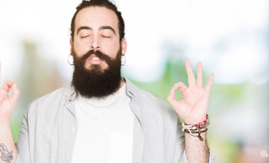 Uzun saç, sakal ve küpeler ile genç adam rahatla ve gülümseyen gözlerle kapalı parmak jestle meditasyon yapıyor. Yoga kavramı.