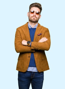 Çapraz kolları olan yüzündeki güneş gözlüğü şüpheci ve sinir, onaylamayan ifade giyen genç yakışıklı zarif adam. Negatif kişi.