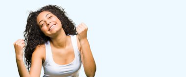 Kıvırcık saçlı kutlama ile genç güzel kız şaşırttı ve başarı için kaldırdı kollarını açarak hayret ve gözleri açık. Kazanan kavramı.