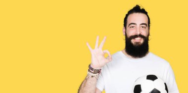 Genç adam uzun saçlı ve parmak, mükemmel sembolü ile ok işareti yapıyor futbol futbol topu tutan ayı