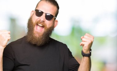 Kutlama güneş gözlüğü takmış genç hipster adam şaşırttı ve başarı için kaldırdı kollarını açarak hayrete ve gözleri açık. Kazanan kavramı.