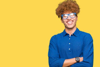 Mavi gözlüklü afro saçlı genç yakışıklı adam kameraya bakarak çapraz kollarıyla gülümseyen mutlu yüz. Pozitif kişi.