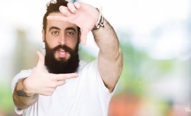 Uzun saç ve sakal elle yapma çerçeve ve parmakları ile mutlu yüz gülümseyen casual beyaz t-shirt giyen genç hipster adam. Yaratıcılık ve Fotoğrafçılık kavramı.