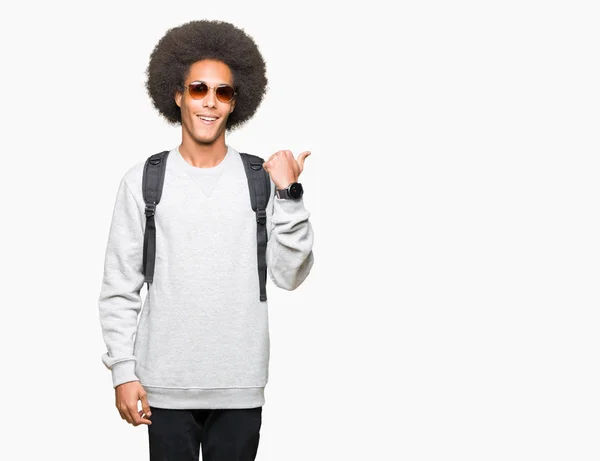 非洲青年美国男子戴着太阳镜和背包 用大拇指指着和快乐的脸微笑着向一边展示 — 图库照片