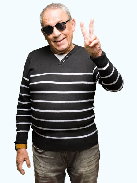 冬のセーターと勝利のサインを行うカメラでウィンクしている幸せそうな顔を浮かべてサングラスを身に着けているハンサムな年配の男性 — ストック写真