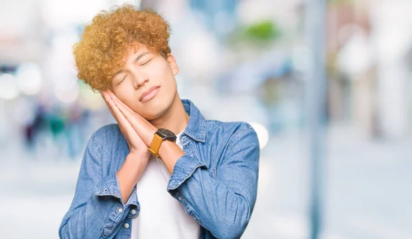 Young handsome man with afro hair wearing denim jacket sleeping tired dreaming and posing with hands together while smiling with closed eyes.