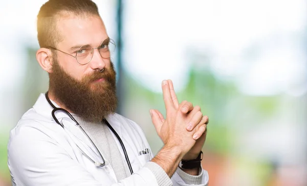 Young blond doctor man with beard wearing medical coat Holding symbolic gun with hand gesture, playing killing shooting weapons, angry face