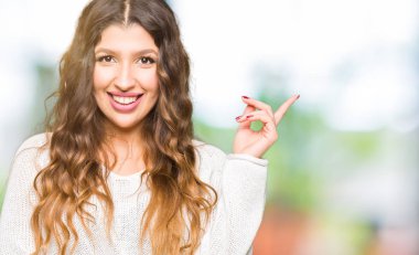 Kameraya bakarak yan el ve parmak ile işaret yüzünde, kocaman bir gülümseme ile beyaz kazak giyen genç güzel kadın.