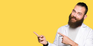 Genç sarışın bilim adam Beyaz önlük giyen gülümseyen ve iki elleri ve parmakları ile tarafına işaret kamera bakıyor.